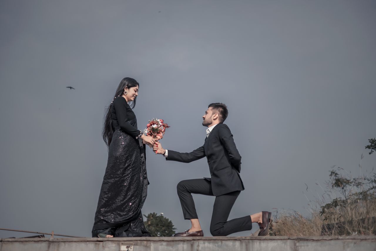 Man proposing with a bouquet, kneeling before a woman in a black dress outdoors.