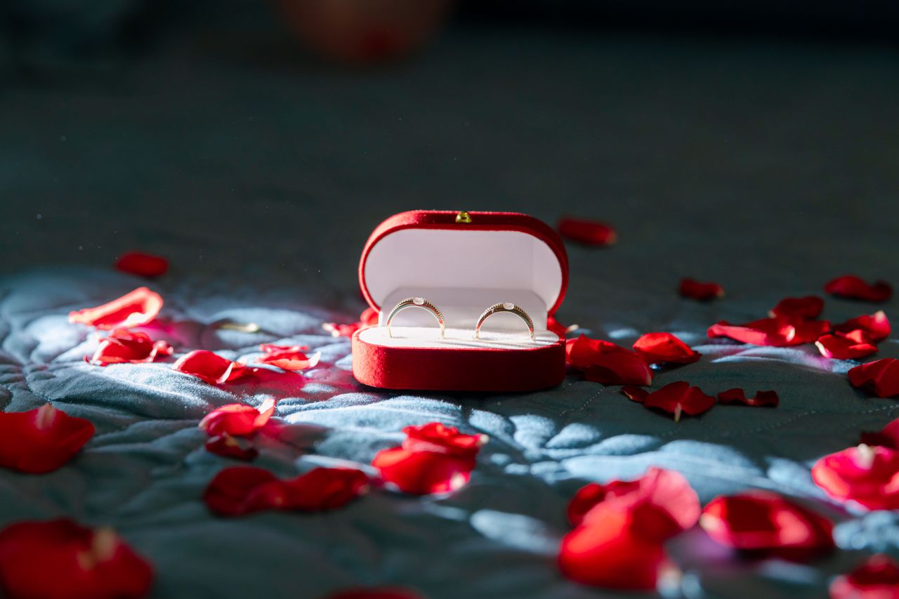 A pair of wedding rings in a red box surrounded by red rose petals on a quilted surface.