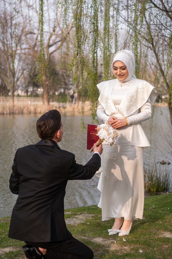 A man proposes to a woman in a white dress by a riverside, capturing a romantic moment.