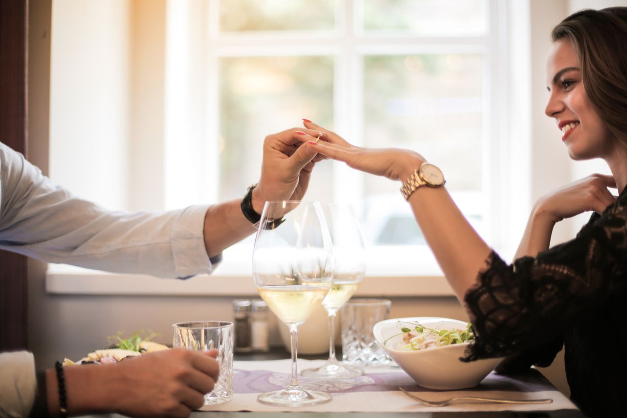 Side view of male putting engagement ring on finger of cheerful elegant female while sitting together at table in restaurant during romantic date and looking at each other